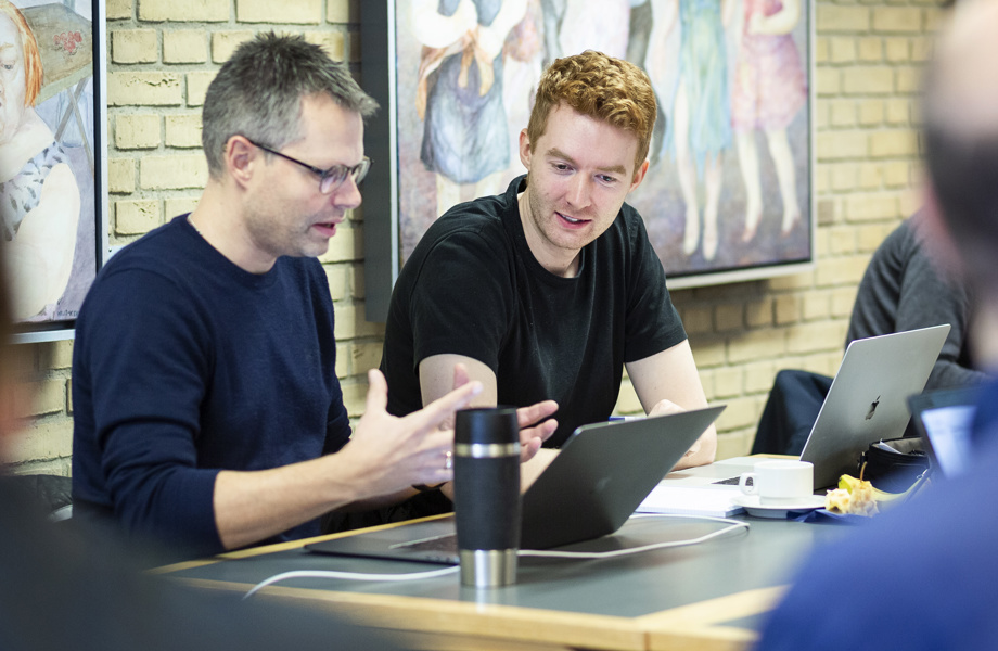 To mænd sidder og laver gruppearbejde sammen på en uddannelsesinstitution. Mændene sidder og snakker om noget der foregår på en bærbar, som er placeret foran dem.