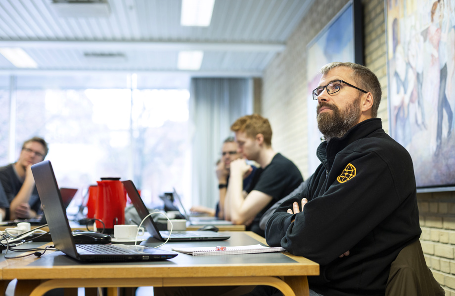 Mand med sort trøje sidder til undervisning. Manden har armene krydset over brystkassen og kigger lyttende op