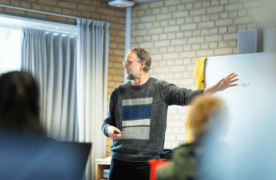 Underviser står ved tavle i et klasselokale. Underviseren er i gang med at holde et oplæg. 